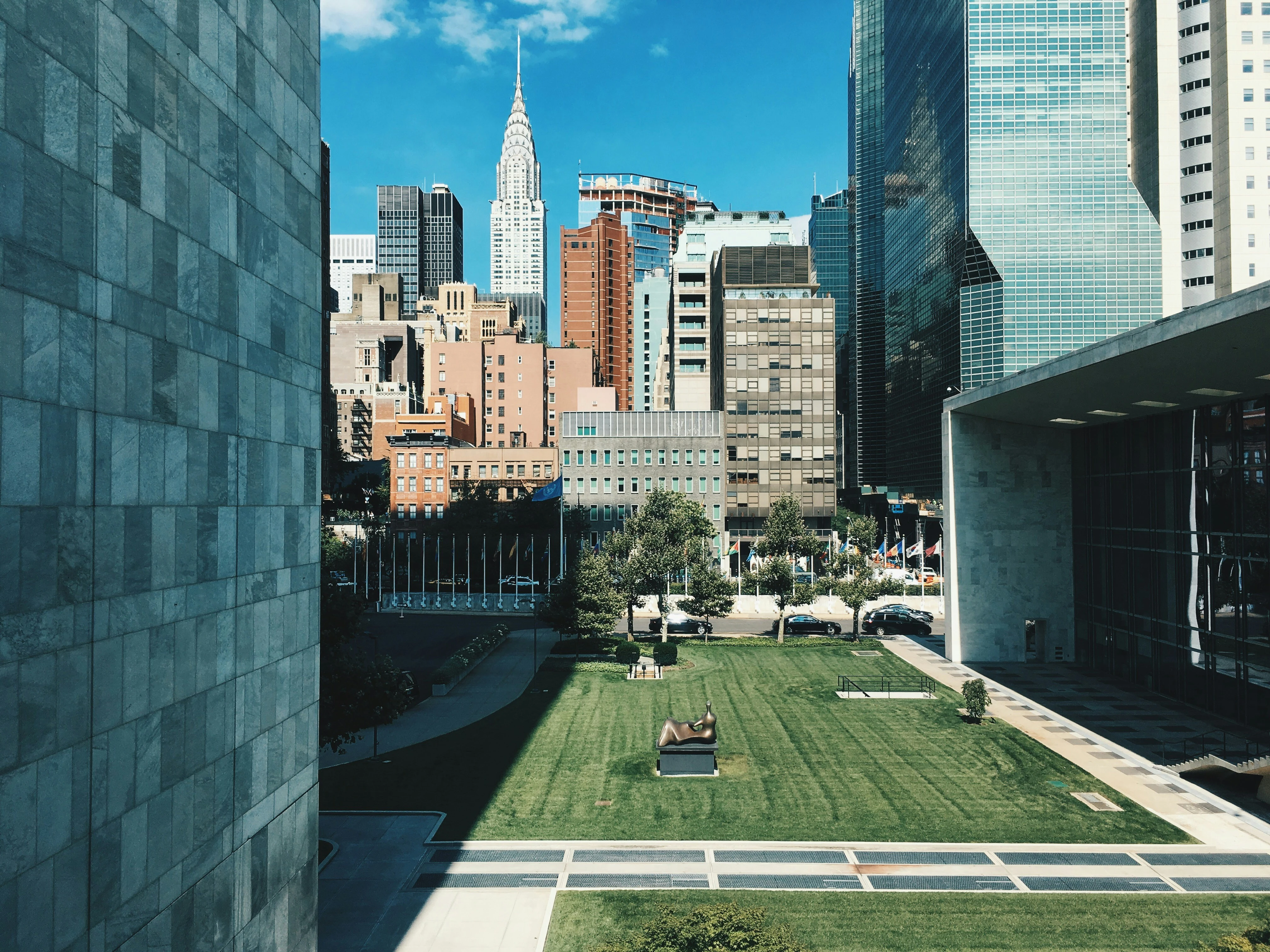 View of the United Nations Headquarters lawn in NYC, with modern buildings on either side and the Chrysler Building visible in the background under a clear blue sky.